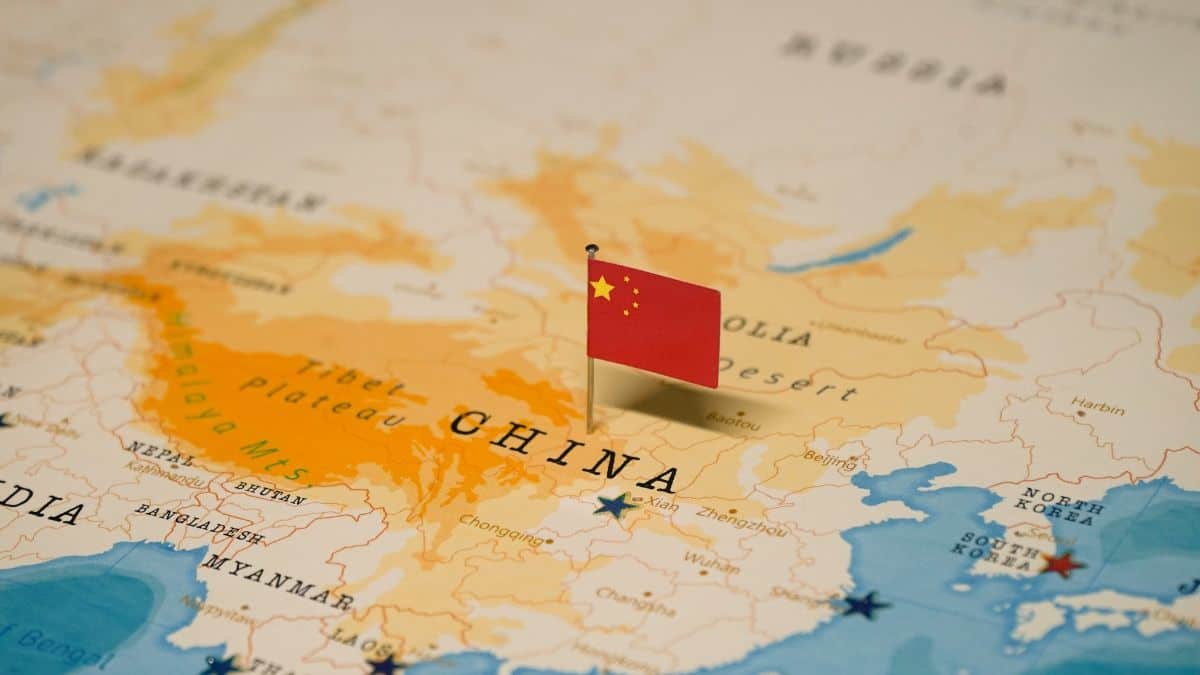 Map of China with a small Chinese flag pinned to mark a location on the country outline and surrounding regions nearby in orange and blue seas.