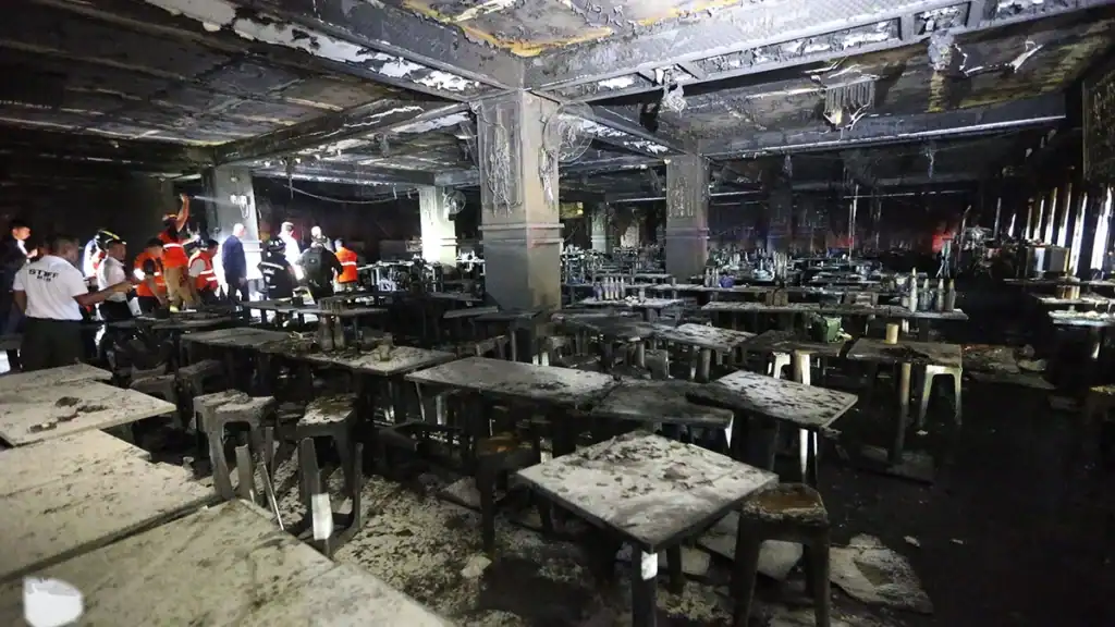 Charred cafeteria interior with ruined tables and stools; search-and-rescue workers in orange vests inspecting the scene.