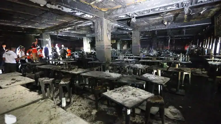 Charred cafeteria interior with ruined tables and stools; search-and-rescue workers in orange vests inspecting the scene.