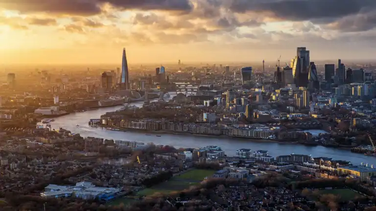 Aerial view of London at sunset, with the River Thames winding through skyscrapers including The Shard and iconic towers inland under a golden sky.