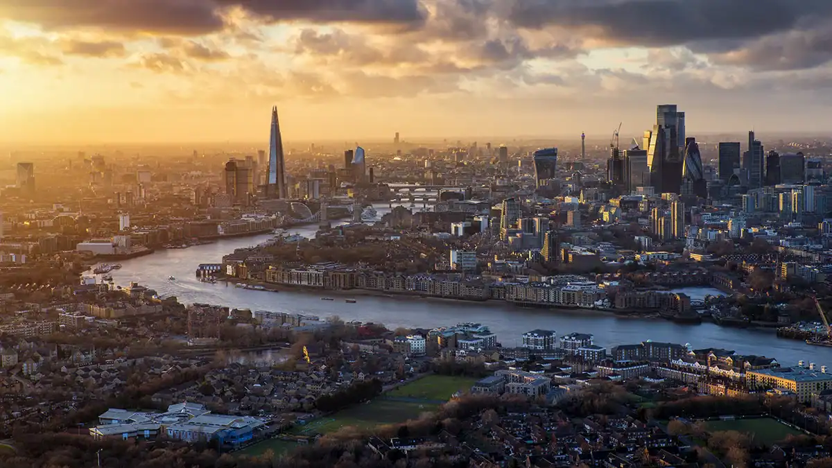 Aerial view of London at sunset, with the River Thames winding through skyscrapers including The Shard and iconic towers inland under a golden sky.