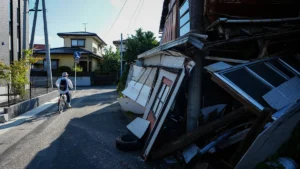 Cyclist wearing a hat rides along a sunny street past a collapsed wooden building with rubble on the right .