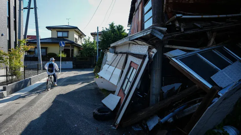 Cyclist wearing a hat rides along a sunny street past a collapsed wooden building with rubble on the right .