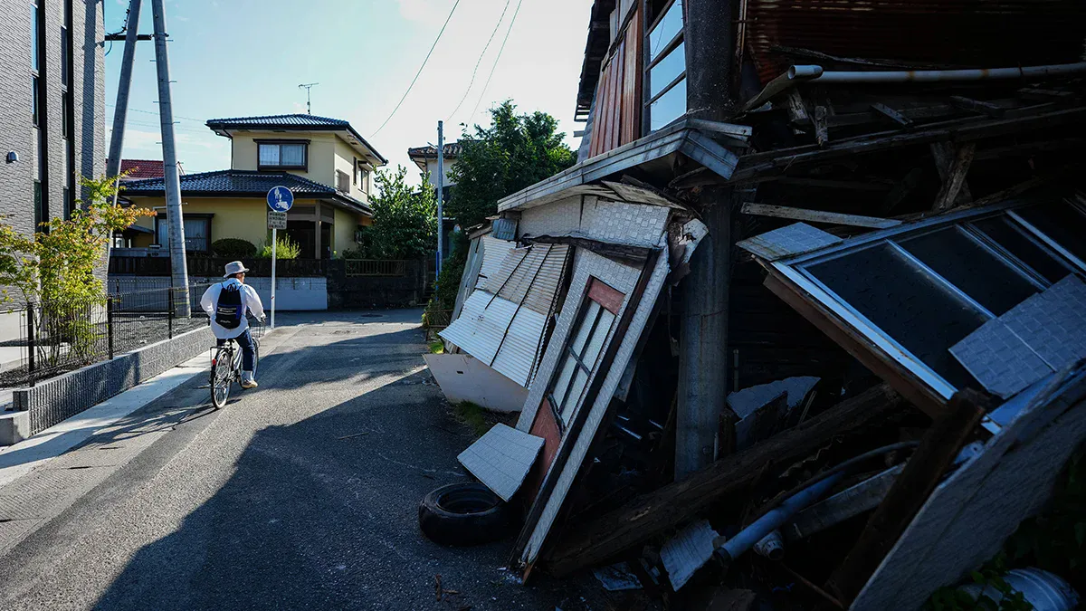 Cyclist wearing a hat rides along a sunny street past a collapsed wooden building with rubble on the right .