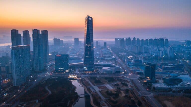 Aerial view of a modern city skyline at sunset with a tall central skyscraper and a river in the distance,