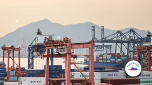 Port scene at sunset with red gantry cranes, stacked shipping containers, and a container ship; mountains in the distance.