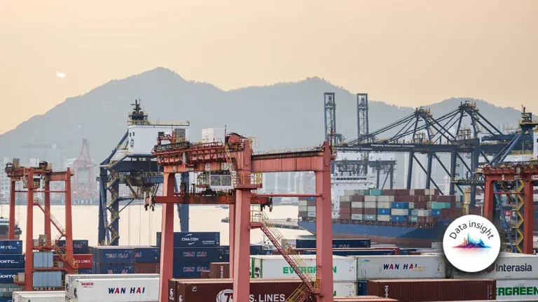 Port scene at sunset with red gantry cranes, stacked shipping containers, and a container ship; mountains in the distance.