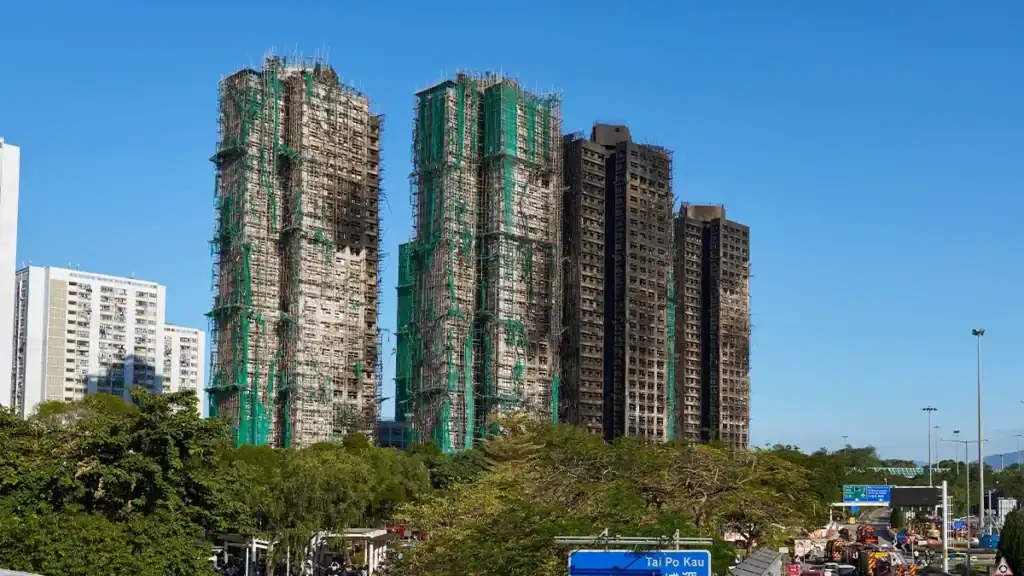 Tall apartment towers under construction wrapped in green netting, with trees and a blue sky in front.
