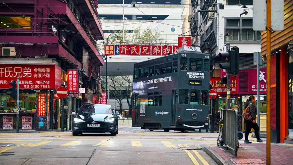 Green double‑decker tram with American Express branding on a busy Chinatown street, surrounded by red storefront signs and a black car ahead.