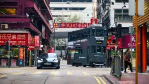 Green double‑decker tram with American Express branding on a busy Chinatown street, surrounded by red storefront signs and a black car ahead.