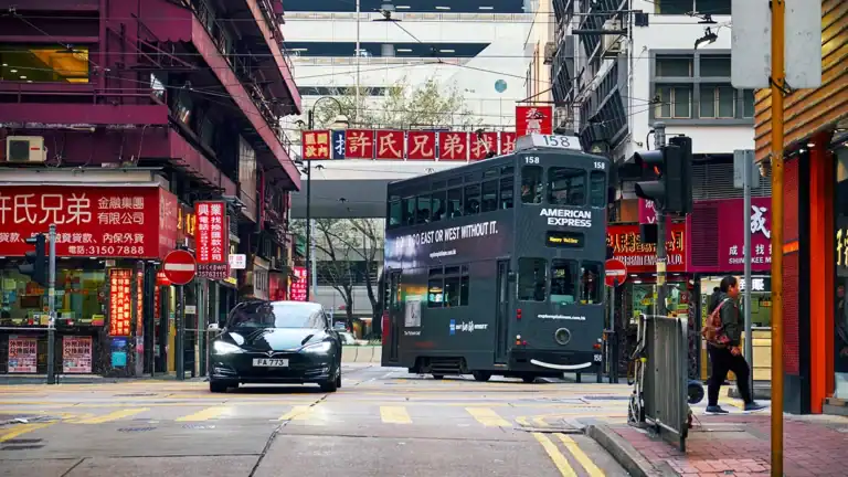 Green double‑decker tram with American Express branding on a busy Chinatown street, surrounded by red storefront signs and a black car ahead.