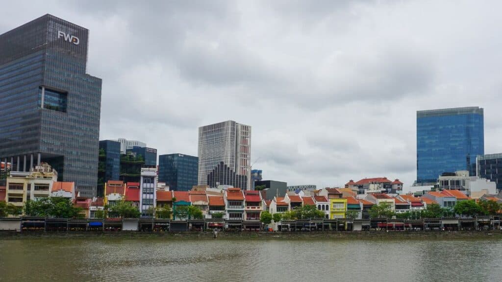 Waterfront row of colorful shophouses with modern glass skyscrapers behind and a cloudy sky.