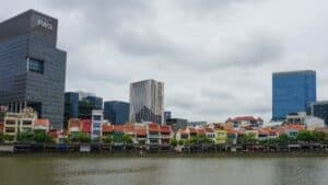 Waterfront row of colorful shophouses with modern glass skyscrapers behind and a cloudy sky.