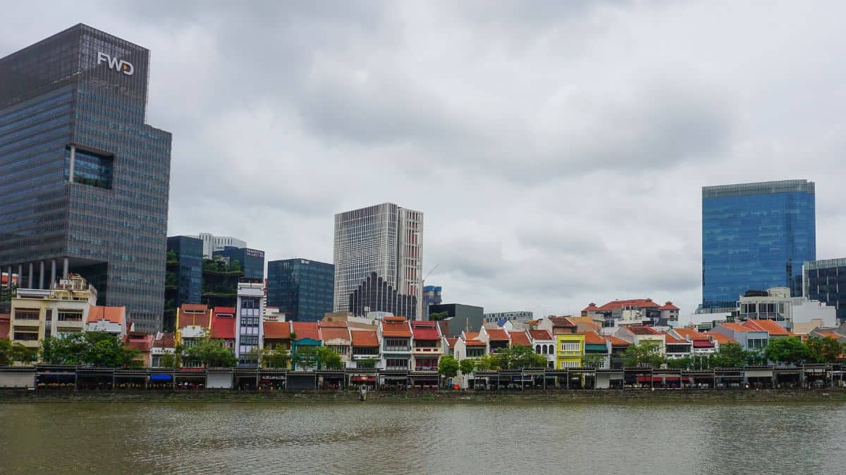 Waterfront row of colorful shophouses with modern glass skyscrapers behind and a cloudy sky.