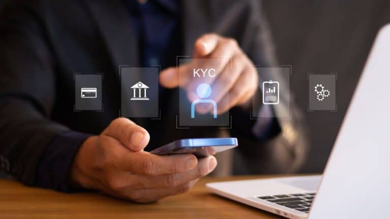 Hand holding a smartphone with floating KYC and banking icons above the screen on a desk setup.