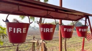 Row of four red metal buckets labeled FIRE hanging from a wooden beam outdoors with a rural field in the background.