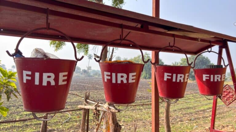 Row of four red metal buckets labeled FIRE hanging from a wooden beam outdoors with a rural field in the background.