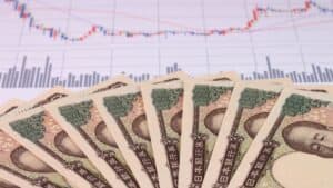 Fanned Japanese yen banknotes in the foreground with a financial stock chart in the background showing price trends and bars in blue and red terms.
