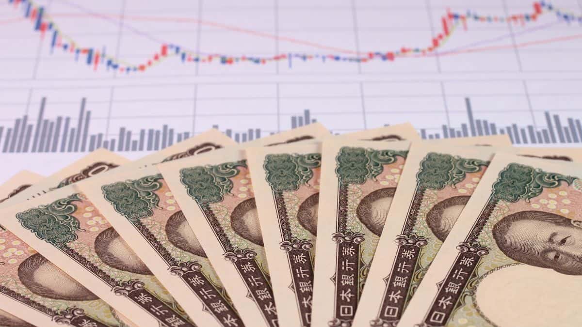 Fanned Japanese yen banknotes in the foreground with a financial stock chart in the background showing price trends and bars in blue and red terms.