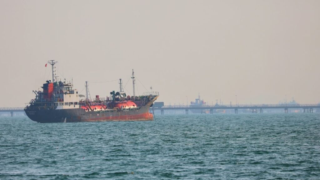 Red and black tugboat with equipment on deck sails in calm sea, a distant pier visible on the horizon.