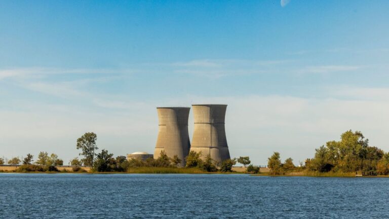 Two large cooling towers rise beside a body of water, with trees and a clear blue sky in the background.