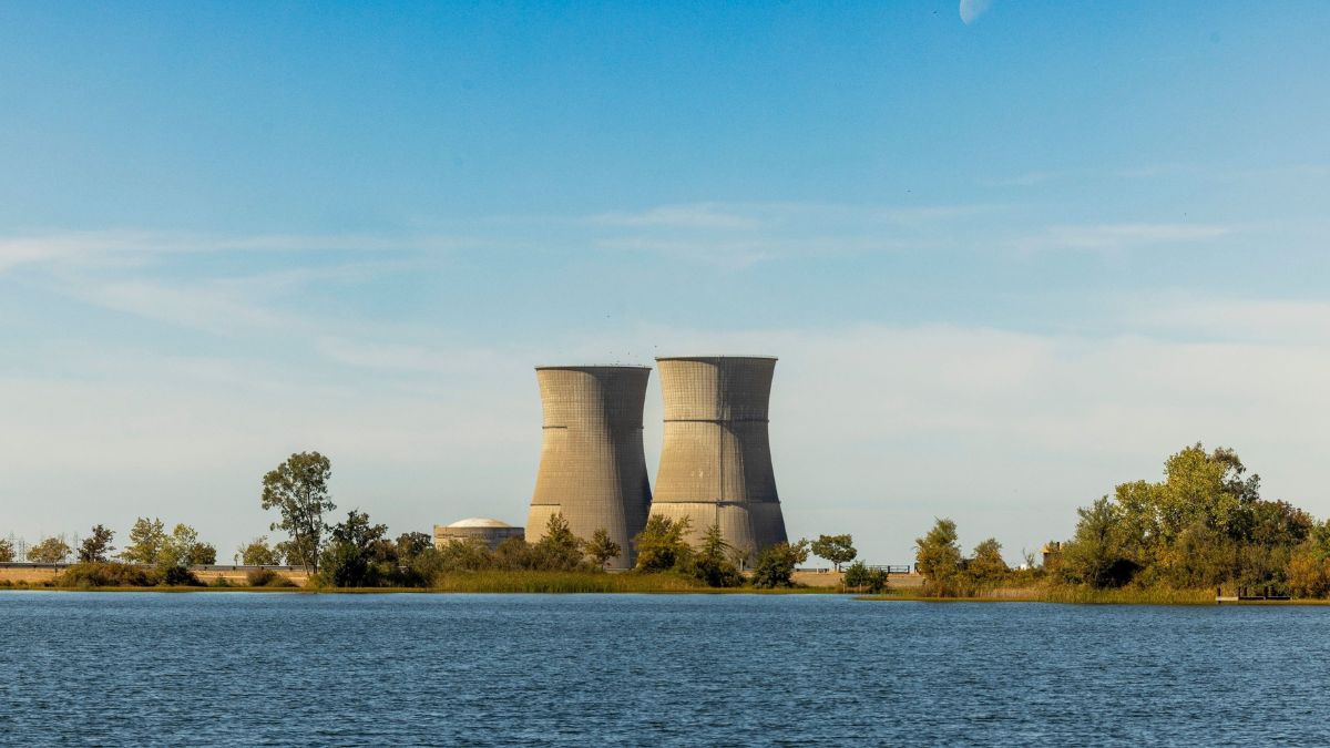 Two large cooling towers rise beside a body of water, with trees and a clear blue sky in the background.