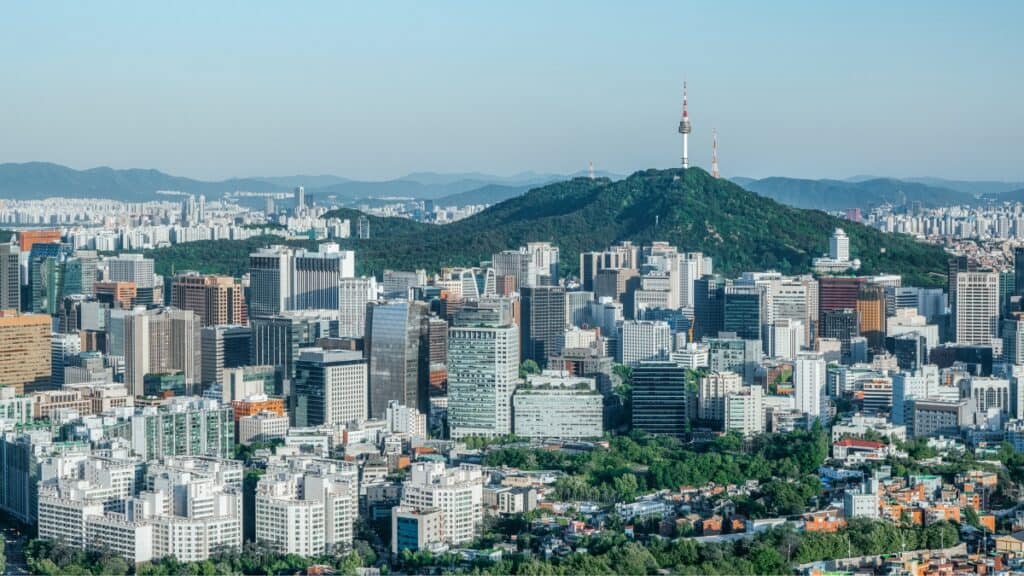 Dense city skyline with a green hill and a tall communications tower rising above the hills in the distance.