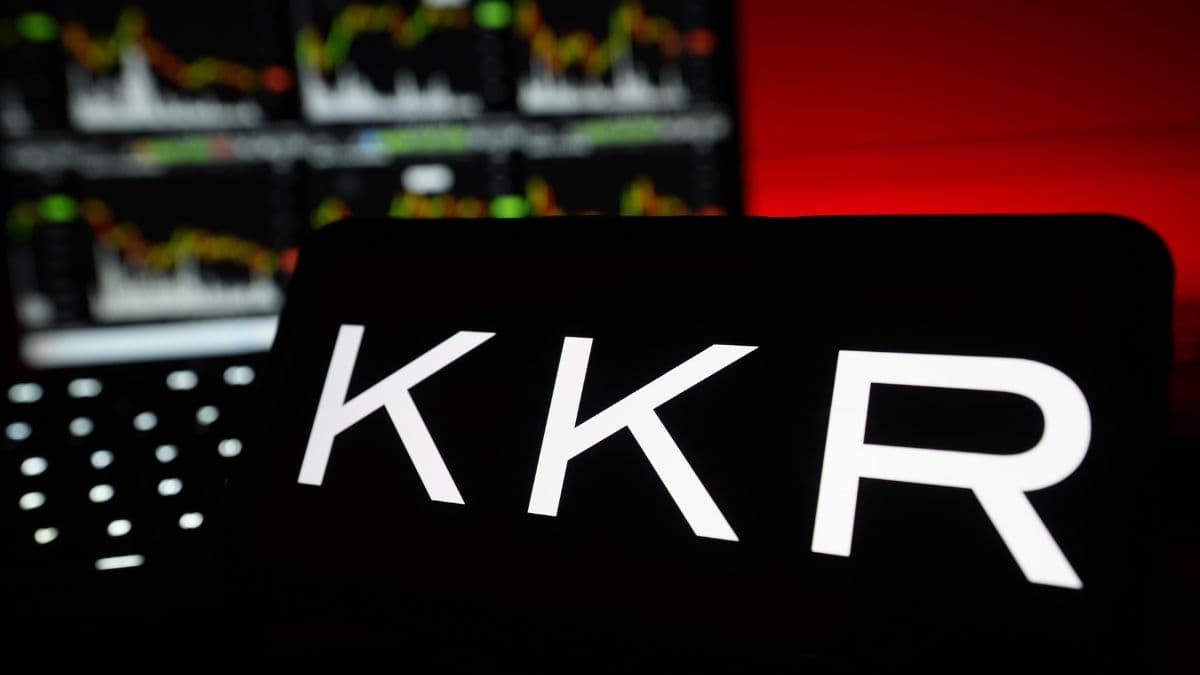 Sign with 'KKR' logo illuminated in a dark room, with blurred stock charts on monitors in the background.