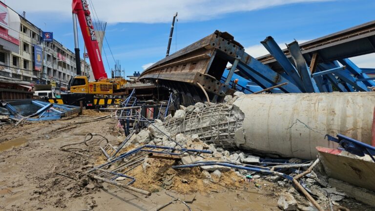 Construction site with heavy equipment and tangled steel amid collapsed concrete rubble on muddy ground and blue sky above.
