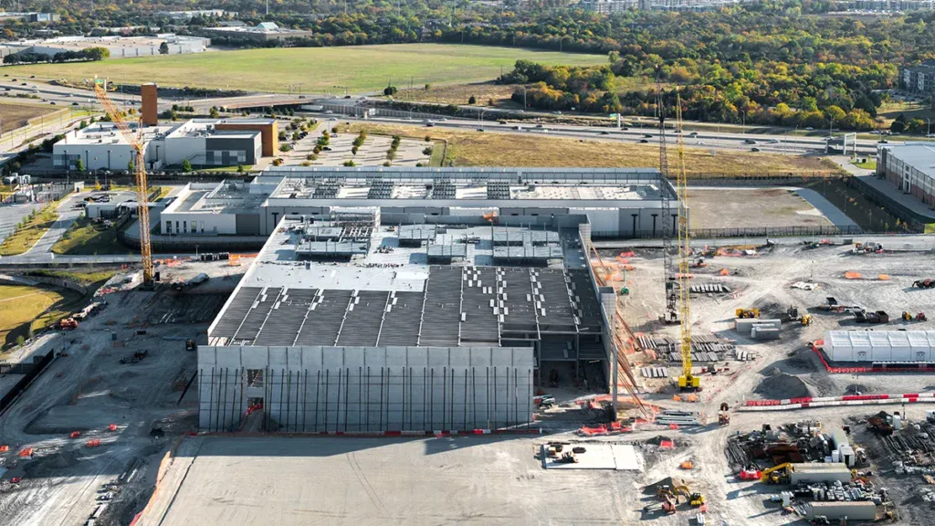 Aerial view of a large construction site with two cranes, a partially built multi-story building, and scattered machinery across the dirt lot.