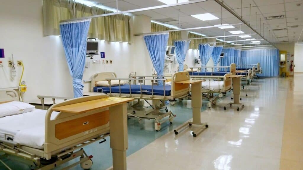 A hospital ward with multiple patient beds separated by blue curtains and medical equipment along the walls under bright ceiling lights.