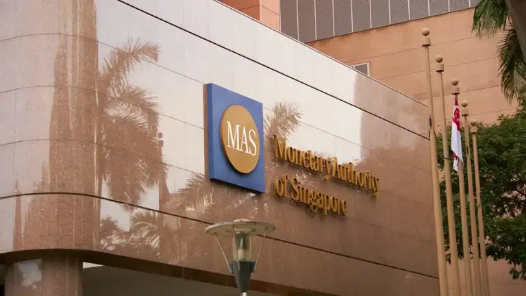 Facade of a building with the Monetary Authority of Singapore sign (MAS logo on blue square) and gold letters reading Monetary Authority of Singapore, with palm trees reflected in the polished wall.