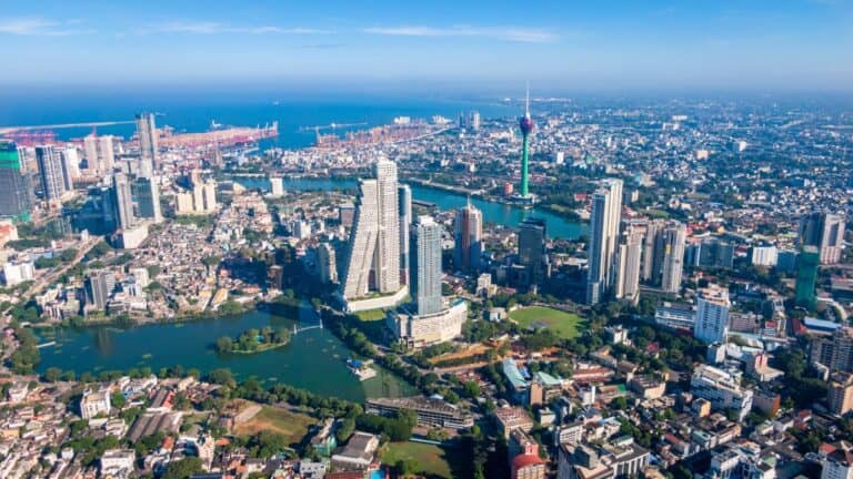 Aerial view of a modern city with high-rise buildings, winding rivers, and a distant harbor under a clear blue sky.",