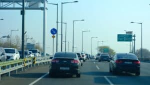 Busy multi-lane highway with cars in both directions; foreground shows a dark sedan beside a metal guard rail and tall streetlights, green Korean road signs ahead.