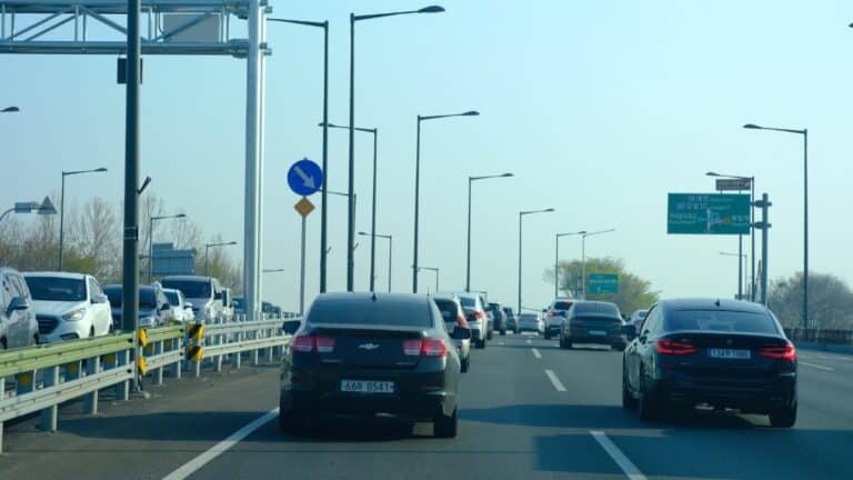 Busy multi-lane highway with cars in both directions; foreground shows a dark sedan beside a metal guard rail and tall streetlights, green Korean road signs ahead.