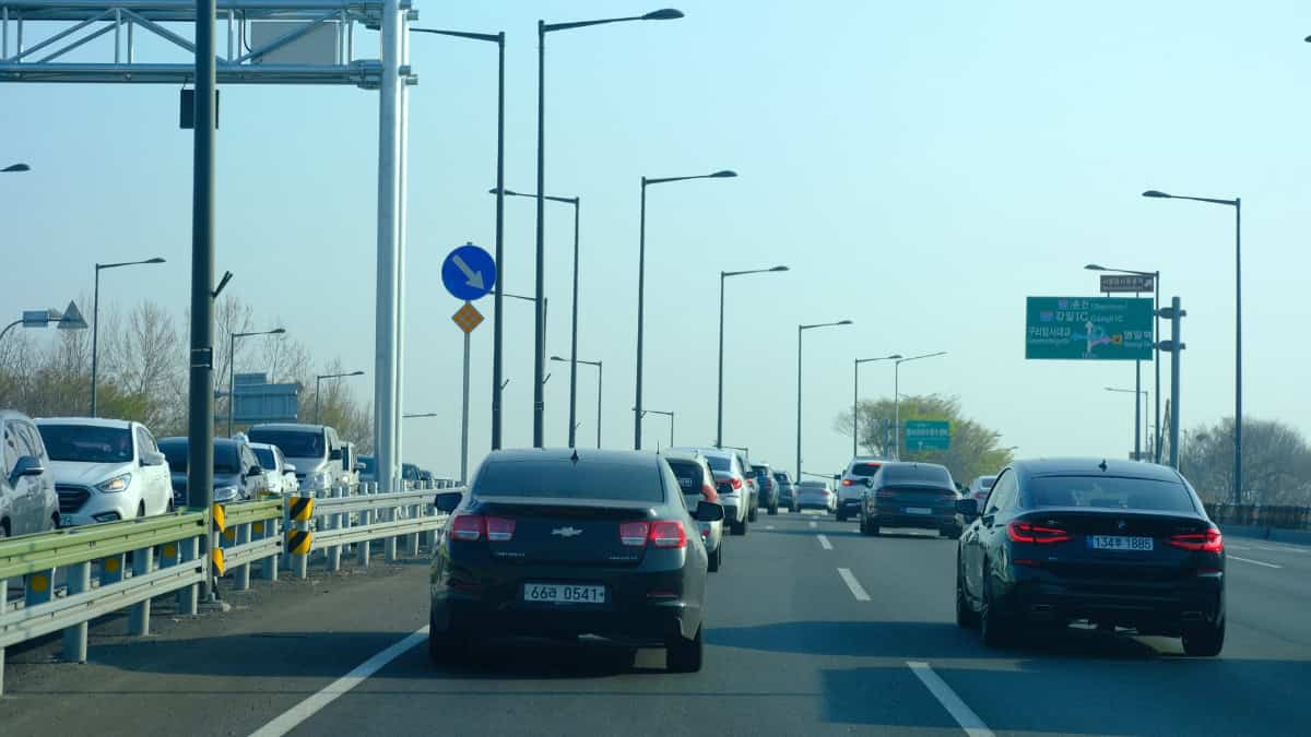 Busy multi-lane highway with cars in both directions; foreground shows a dark sedan beside a metal guard rail and tall streetlights, green Korean road signs ahead.