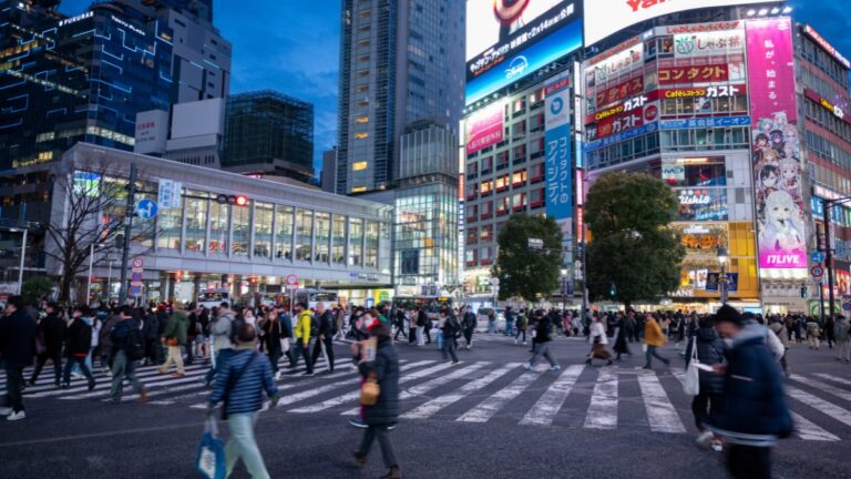 Busy city crosswalk at dusk with many pedestrians crossing a zebra-striped crosswalk, surrounded by brightly lit skyscrapers and large billboards.