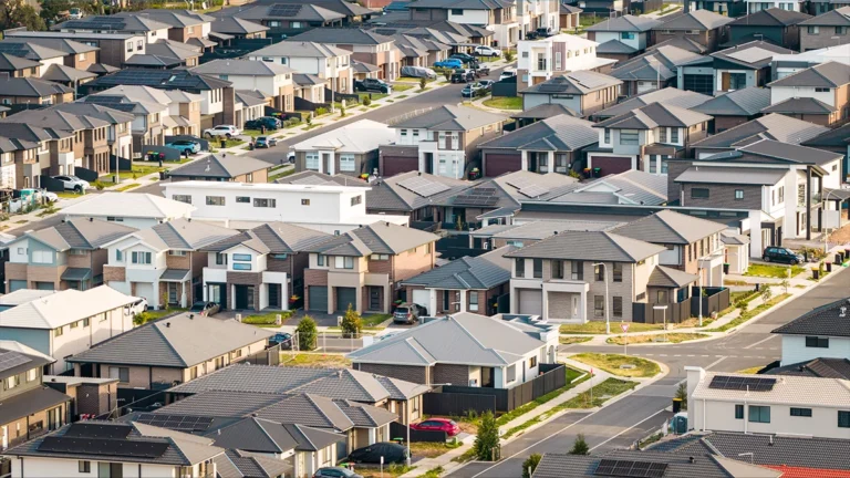 Aerial view of a dense suburban neighborhood with rows of modern two‑story houses and winding streets on a sunny day.