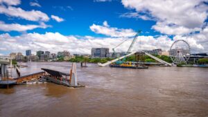 Riverfront cityscape with a large Ferris wheel and a crane-supported structure over the river, under a blue sky with clouds.