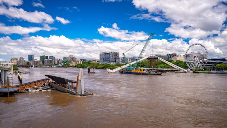 Riverfront cityscape with a large Ferris wheel and a crane-supported structure over the river, under a blue sky with clouds.