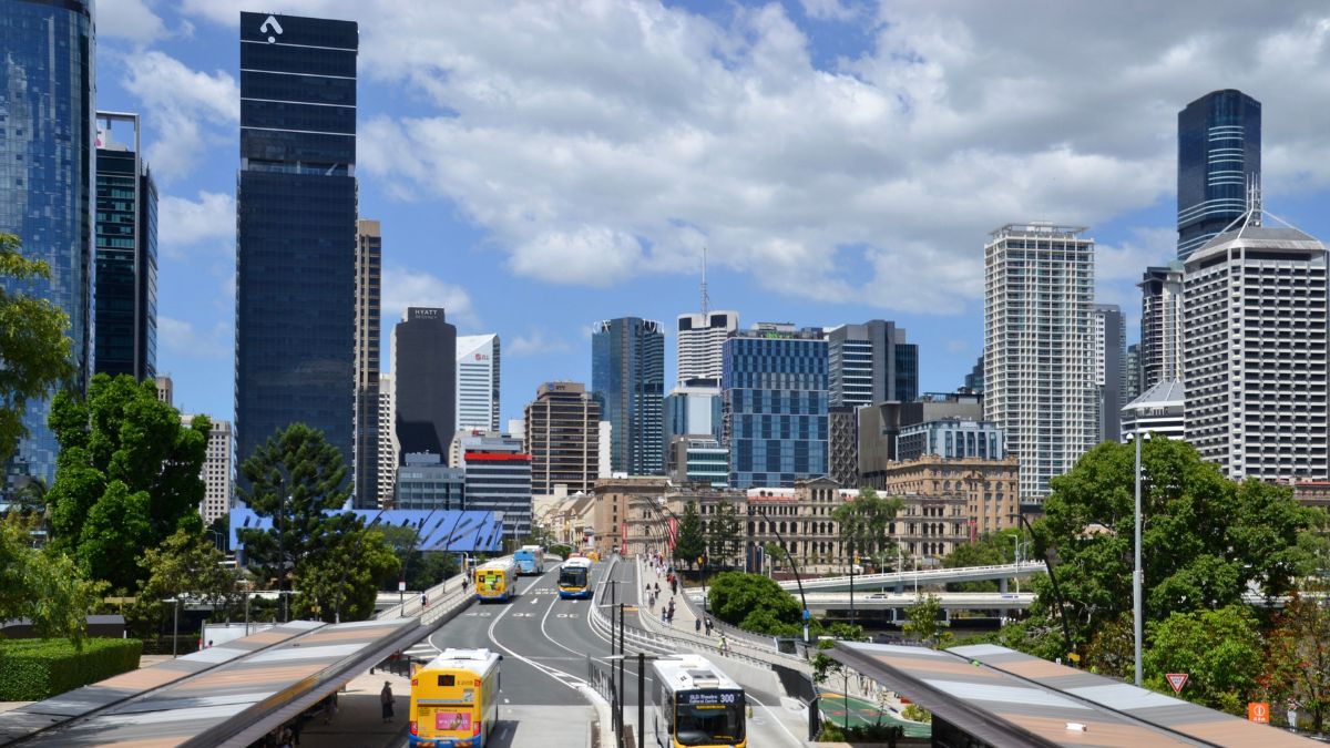 City skyline with tall glass skyscrapers, a multi-lane road, and buses in the foreground.