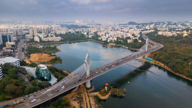 Aerial view of a city with a prominent cable-stayed bridge over a wide river lake surrounded by buildings and green spaces, taken from above the skyline.