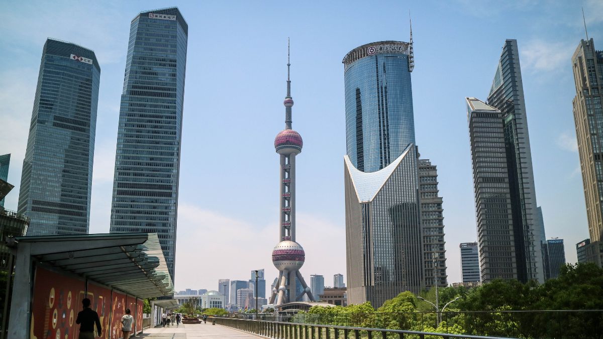 Skyline of Shanghai with the Oriental Pearl Tower, flanked by tall glass skyscrapers on a sunny day, with a pedestrian promenade in the foreground.