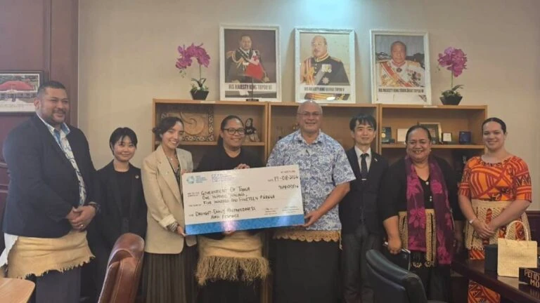 Eight adults posing in a formal office holding a large ceremonial check, smiling for the camera and flanked by colleagues.