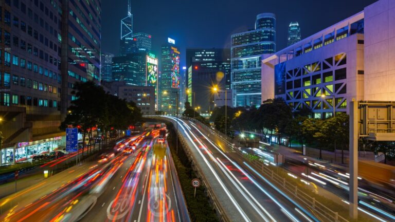 Nighttime city scene with light trails on a busy highway and tall illuminated skyscrapers in the background.