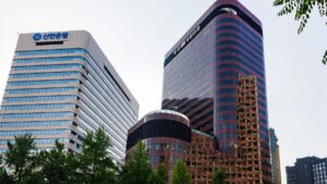 Two modern glass office skyscrapers in a city, left building has a Korean bank sign and trees in the foreground.