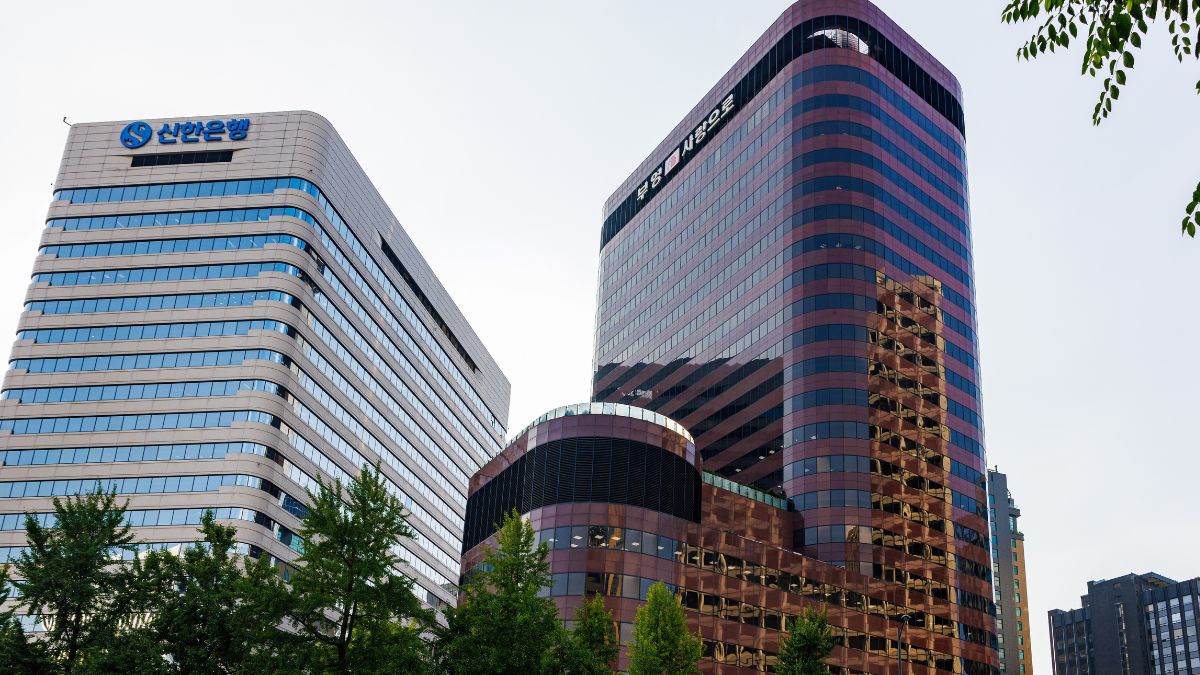 Two modern glass office skyscrapers in a city, left building has a Korean bank sign and trees in the foreground.