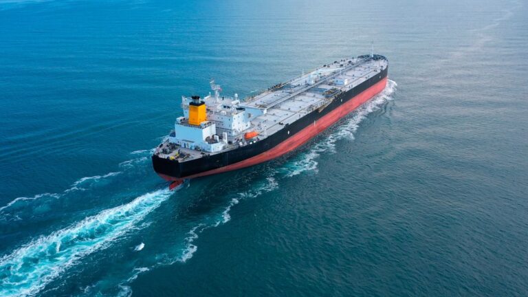 Aerial view of a large black-and-red oil tanker sailing through calm blue sea with a yellow smokestack and white superstructure at the stern.