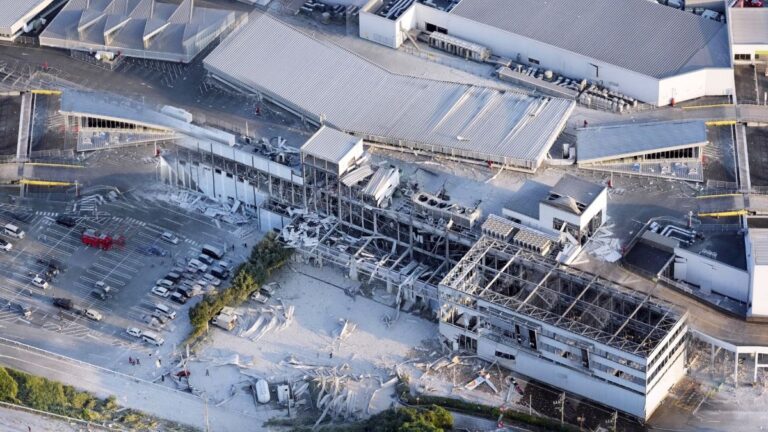Aerial view of a large shopping complex with a partially collapsed building, exposed steel framework, and a debris-filled area nearby.