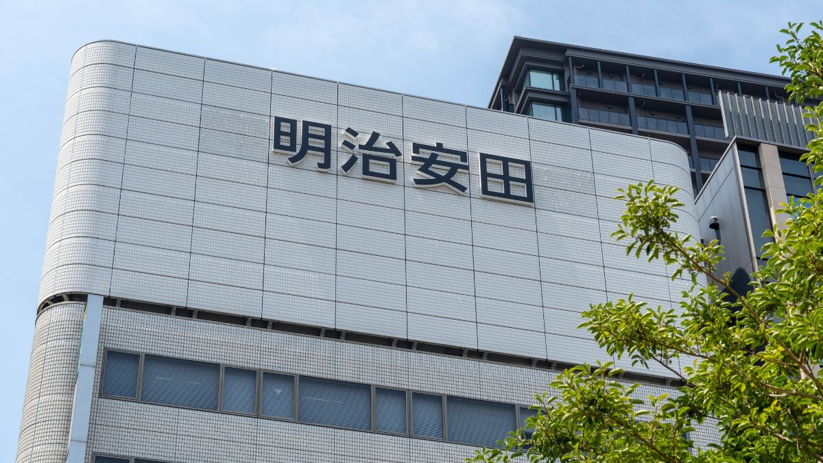 White tiled office building with the Meiji Yasuda sign in Japanese characters, blue sky above, and green trees in the foreground.